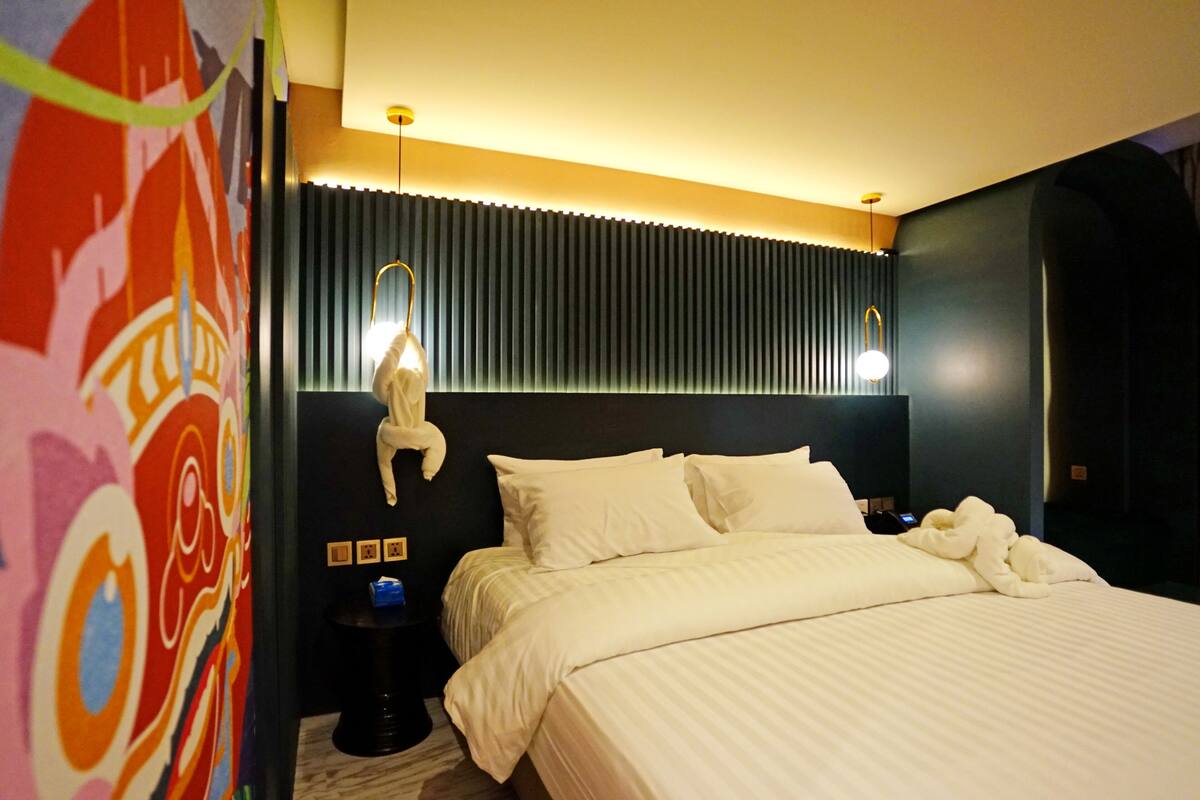 Habitación de hotel con una cama grande, cabecera con listones verticales, lámpara fijada a la pared y un mural abstracto y colorido en la pared izquierda.