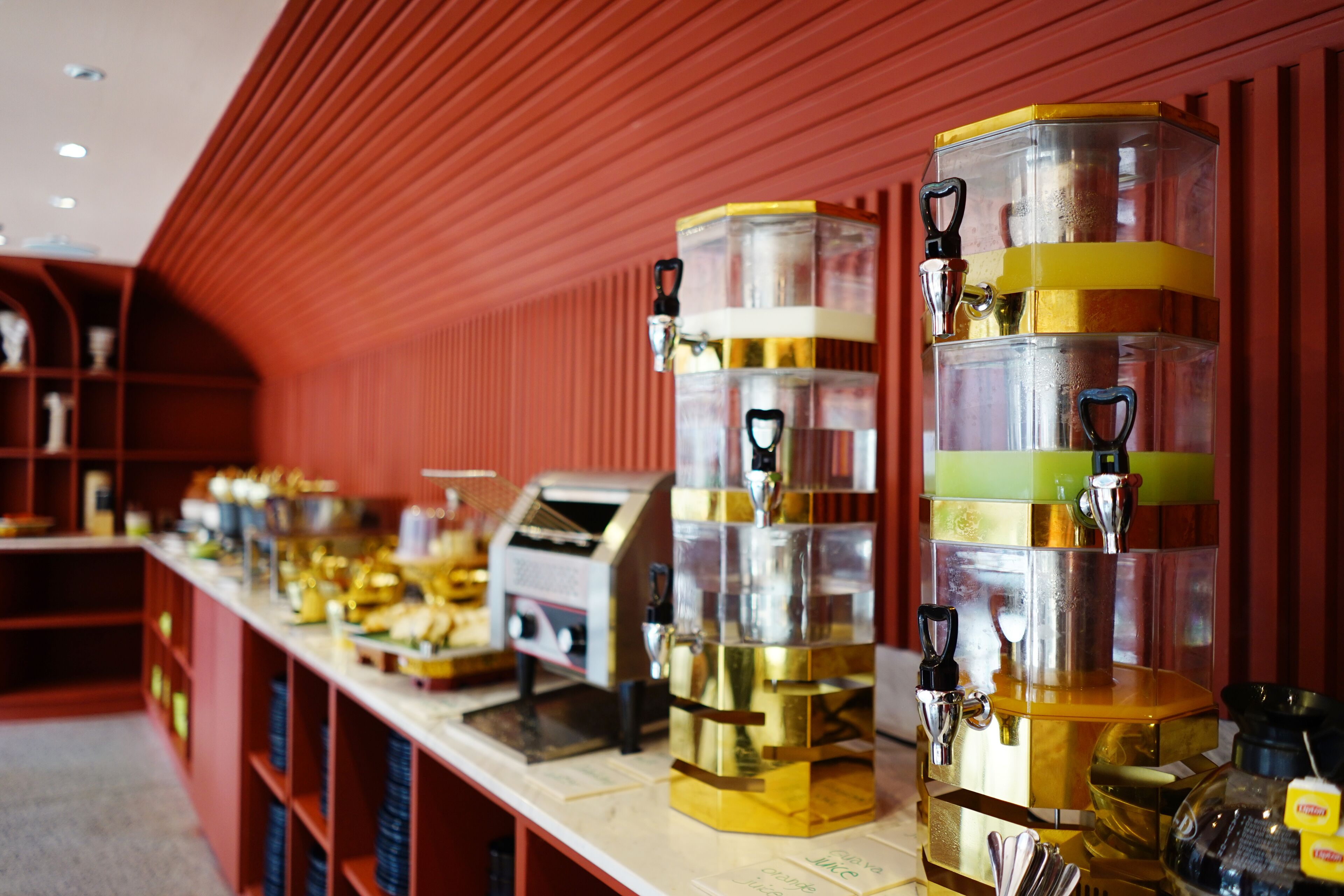 A row of coffee dispensers with multiple spouts, a cash register, and a menu board on a counter in a room with red walls and wooden shelves.