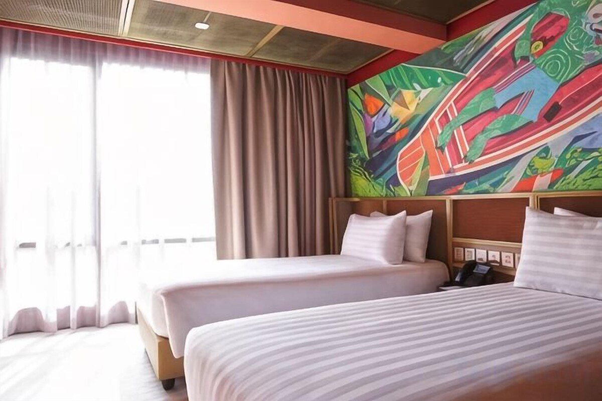 Quarto de hotel com duas camas, uma janela grande com cortinas e um quadro abstrato colorido na parede.