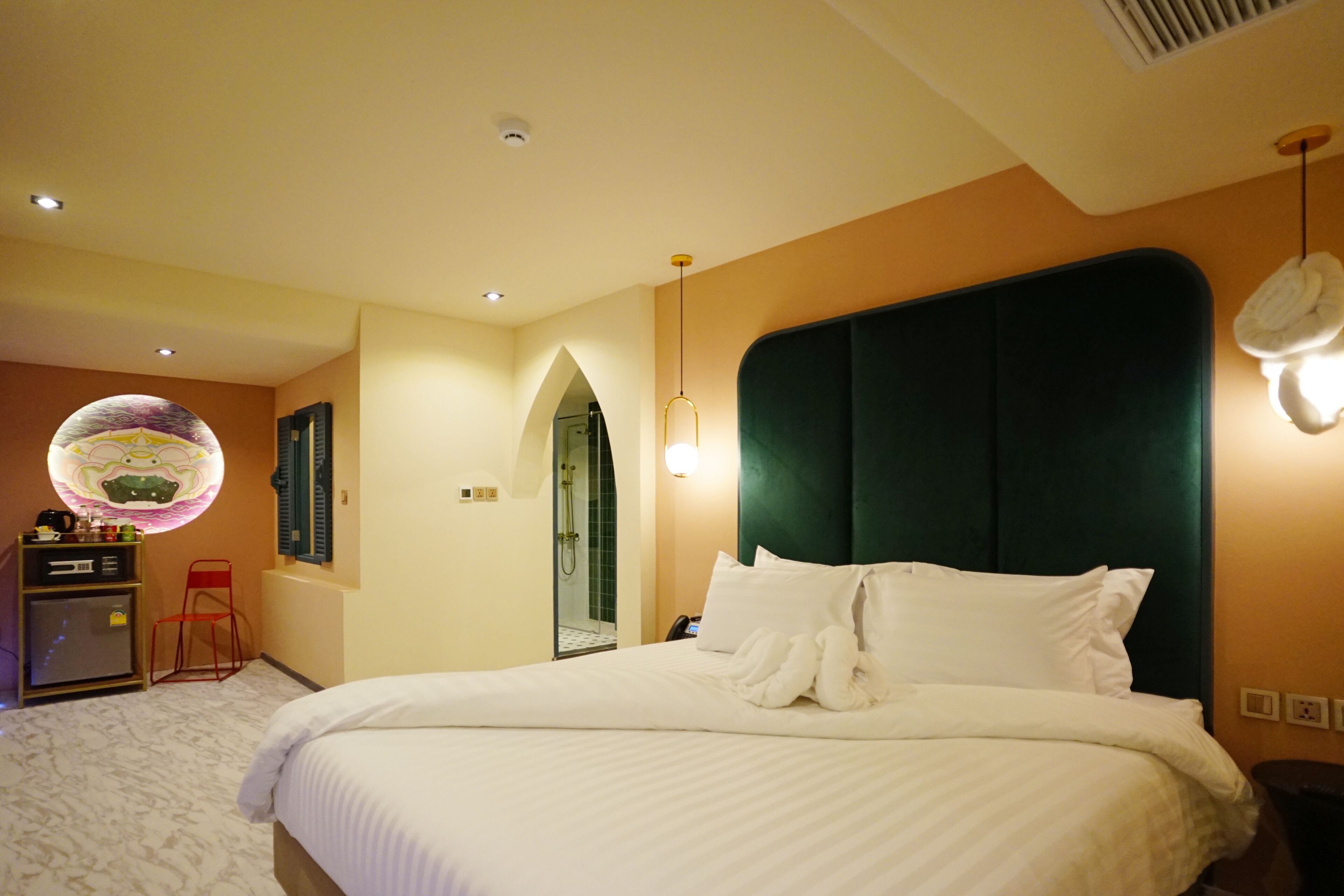 A hotel room with a large bed, a desk with a chair, a mirror, and a circular light fixture.