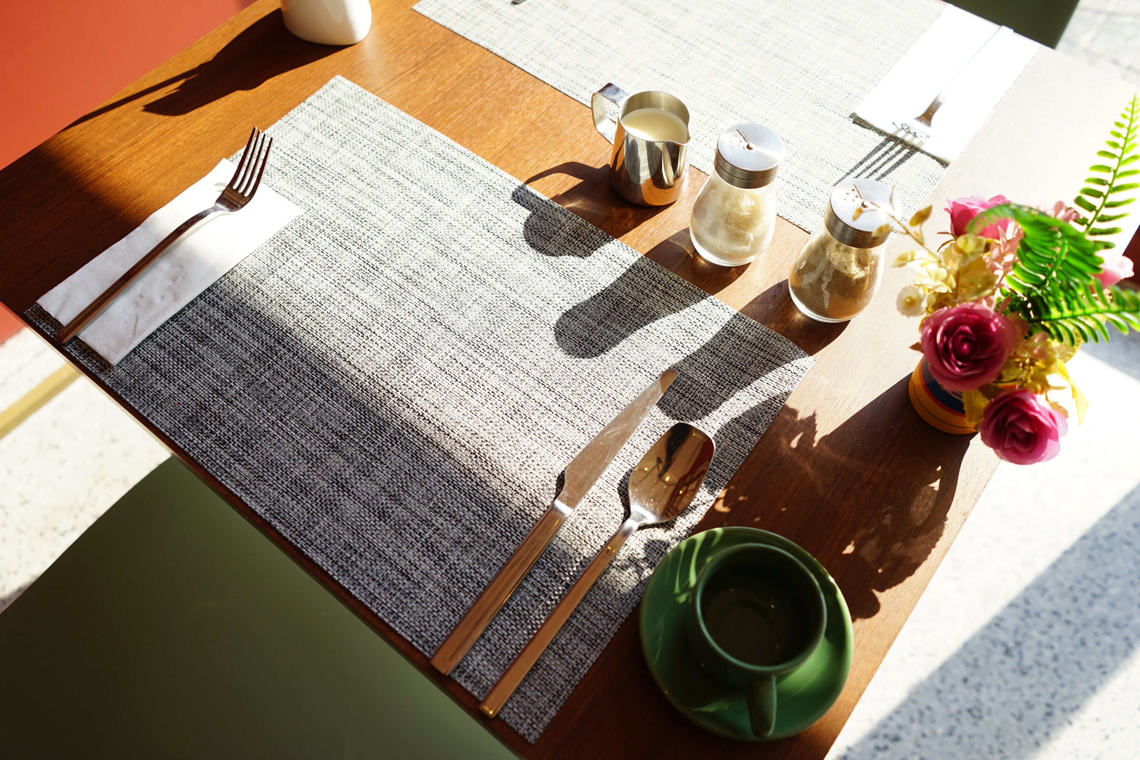 A table set with a placemat, cutlery, a teapot, salt and pepper shakers, a cup, and a vase with flowers.
