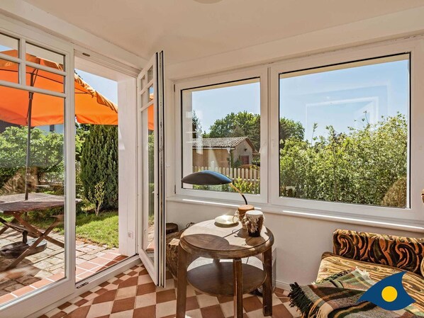 A room with a checkered floor, a round wooden table, a sofa with a patterned throw, and a window with an orange umbrella outside.