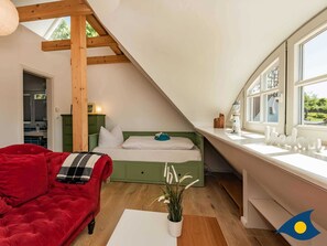 A cozy attic bedroom with a sloped ceiling, a red sofa, a bed with green headboard, and a window with a view of greenery.