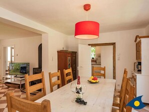 A dining area with a white table, wooden chairs, a red hanging light, and a television in the background.