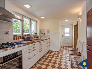 A modern kitchen with a gas stove, a sink, and a window with a view of greenery. There is a checkered floor pattern and a door leading to another room.