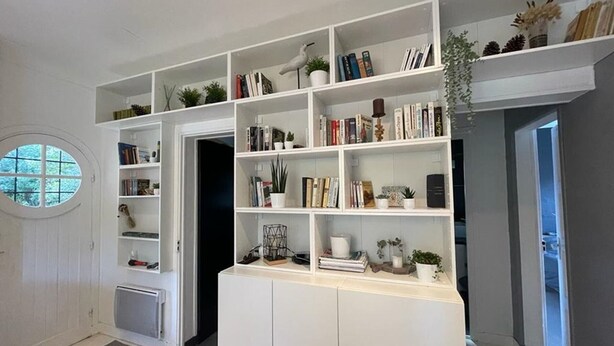 Ein modernes Wohnzimmer mit weißen Wänden und einem großen Fenster. Es verfügt über eine Einbauschränke mit Büchern und dekorativen Gegenständen.