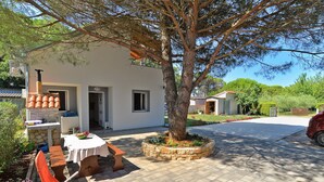 A white building with a stone bench and orange chairs outside, a tree in the center, and a paved driveway leading to a car. - Apartment 1713/17699 (Istria - Valbandon), 200m from the beach (Valbandon)