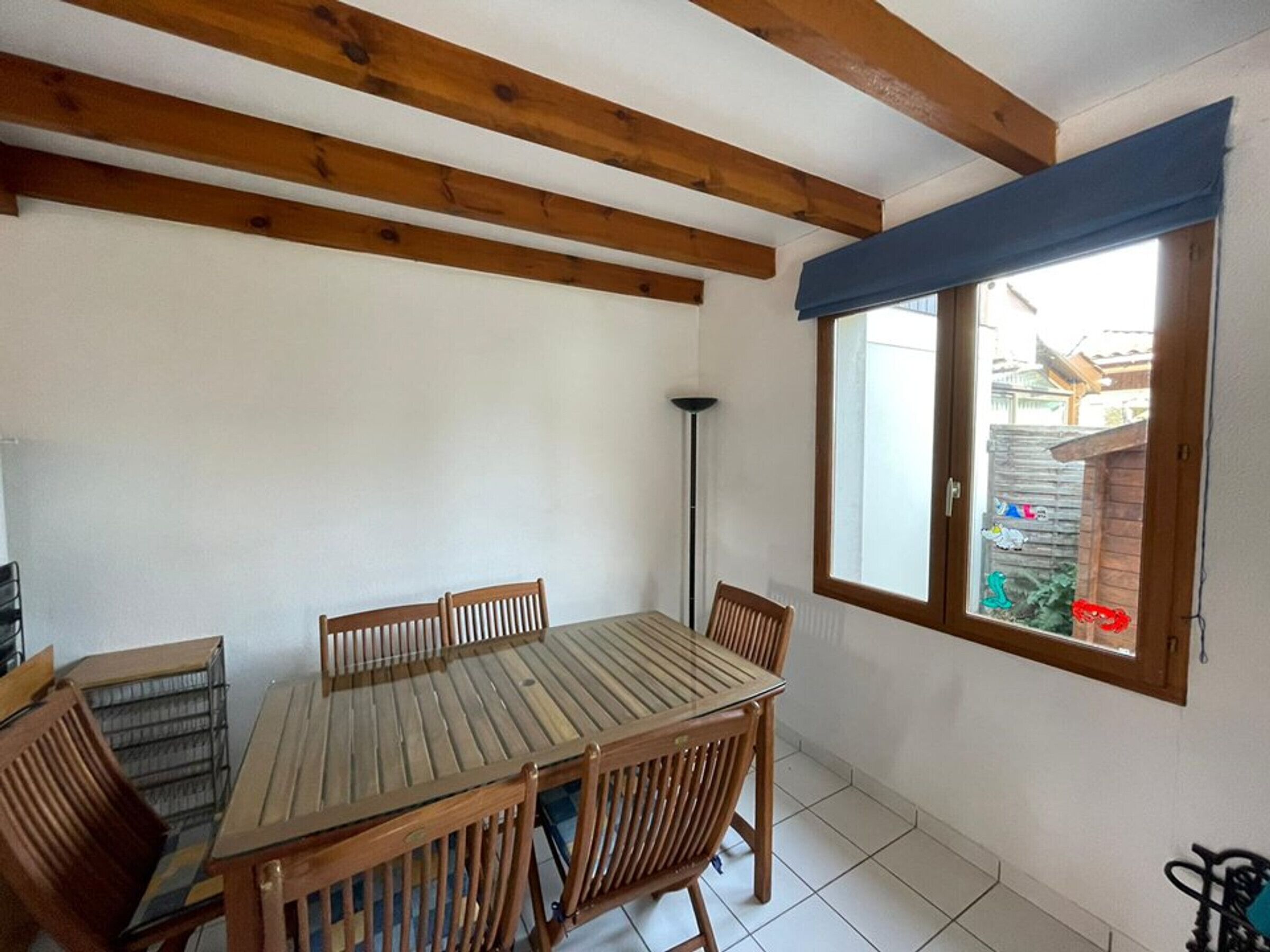 A dining area with a wooden table and chairs, a window with a view of a garden, and a ceiling with exposed beams.