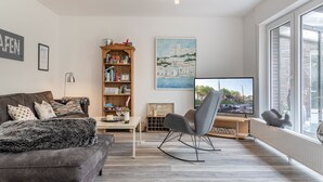 A modern living room with a grey sofa, a wooden bookshelf, a flat-screen TV, and a large window.