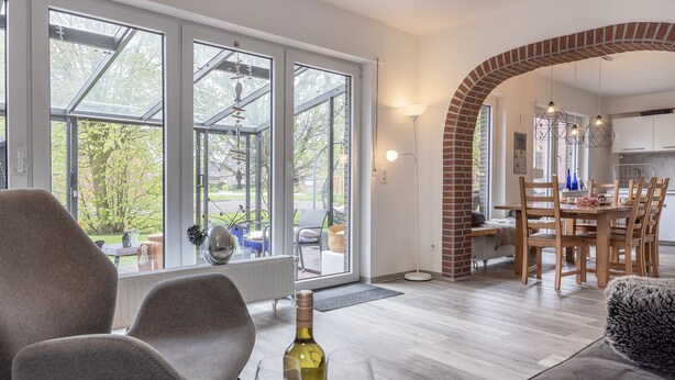 A modern living room with a large window, a dining area with wooden furniture, and a kitchen visible through an archway.