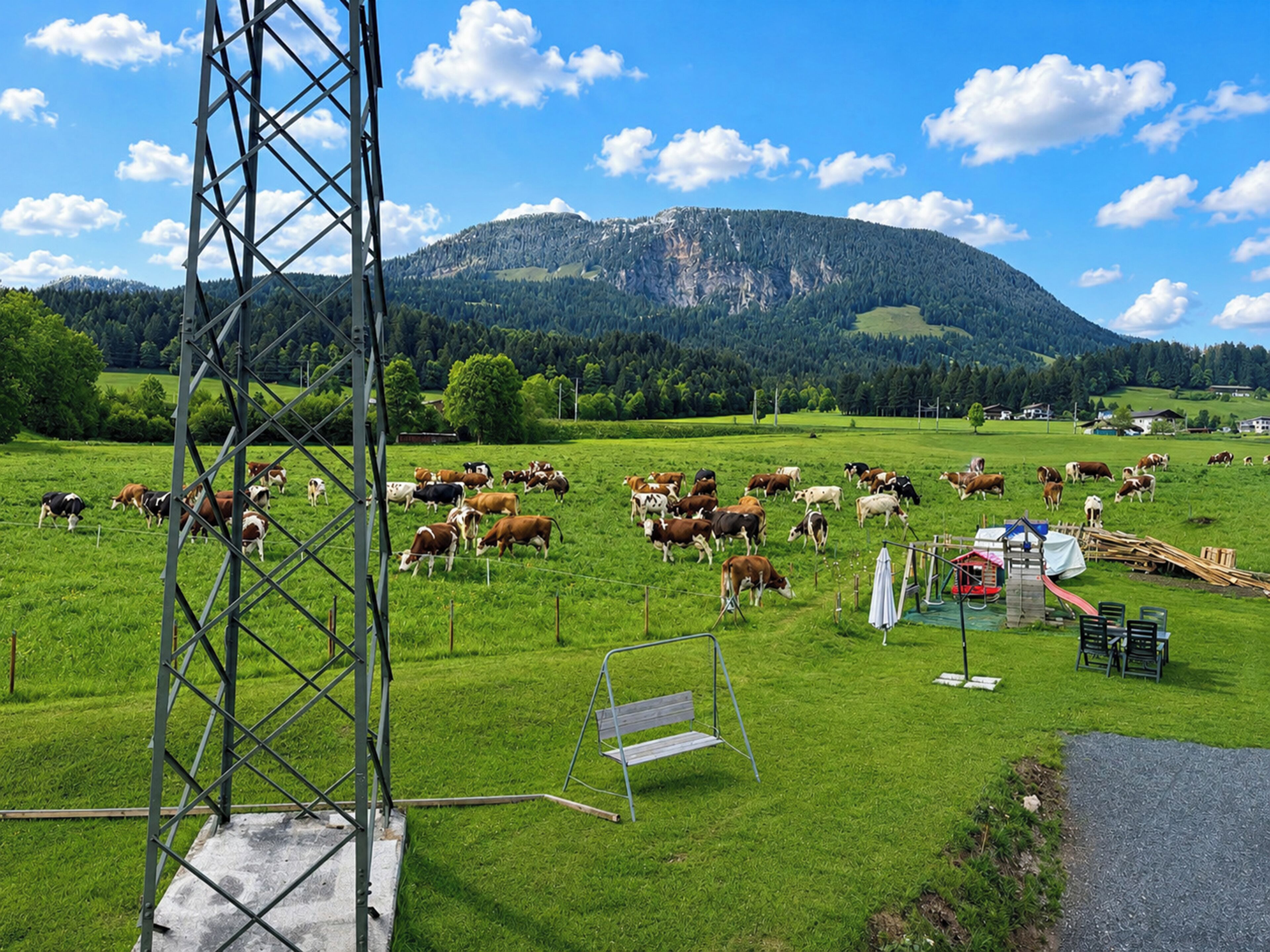 Un paysage rural avec un champ où paissent des vaches, une structure métallique et une montagne en arrière-plan.