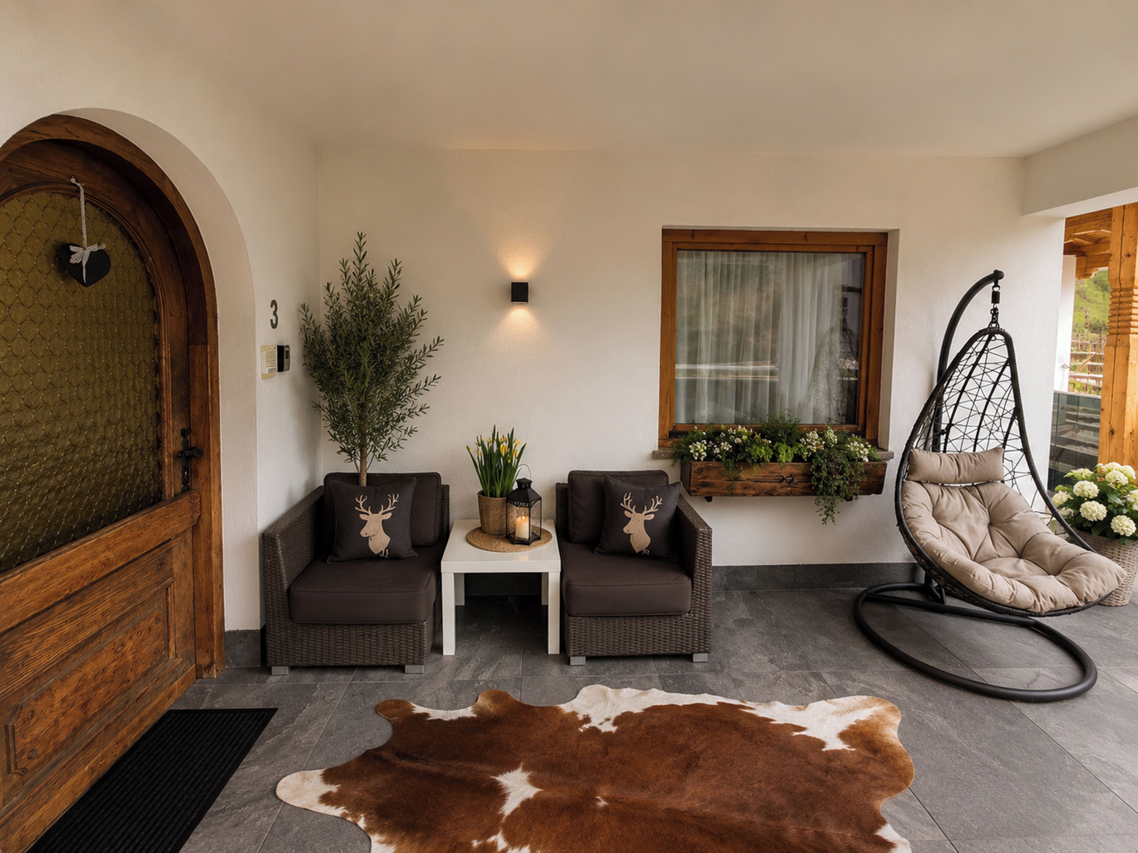 A cozy interior with a wooden door, two sofas, a hanging chair, a small table with a lantern, and a cowhide rug.