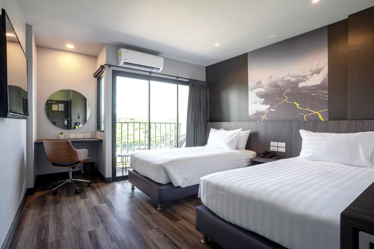 Ein modernes Hotelzimmer mit zwei Betten, einem Schreibtisch, einem Stuhl, einem großen Fenster und einer Wand-Klimaanlage.