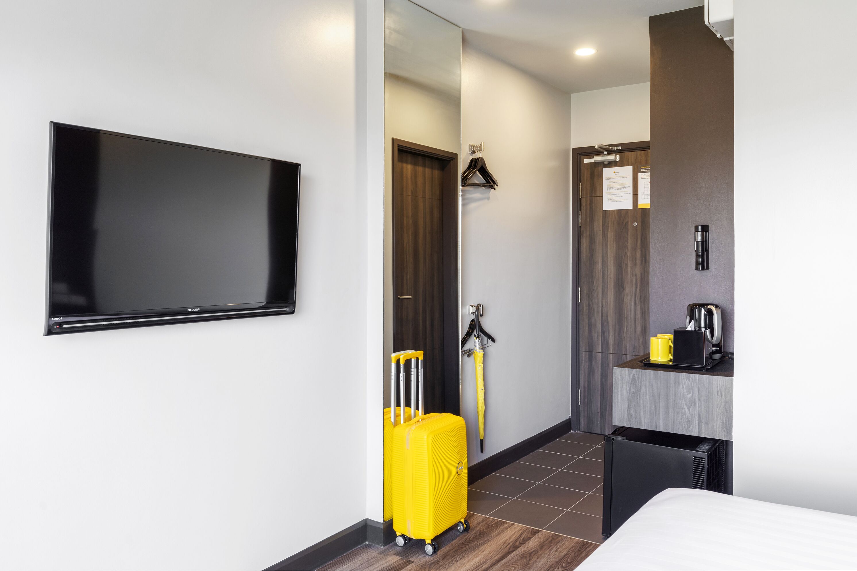 A modern hotel room with a flat-screen TV, a yellow suitcase, a compact refrigerator, and a small coffee maker.