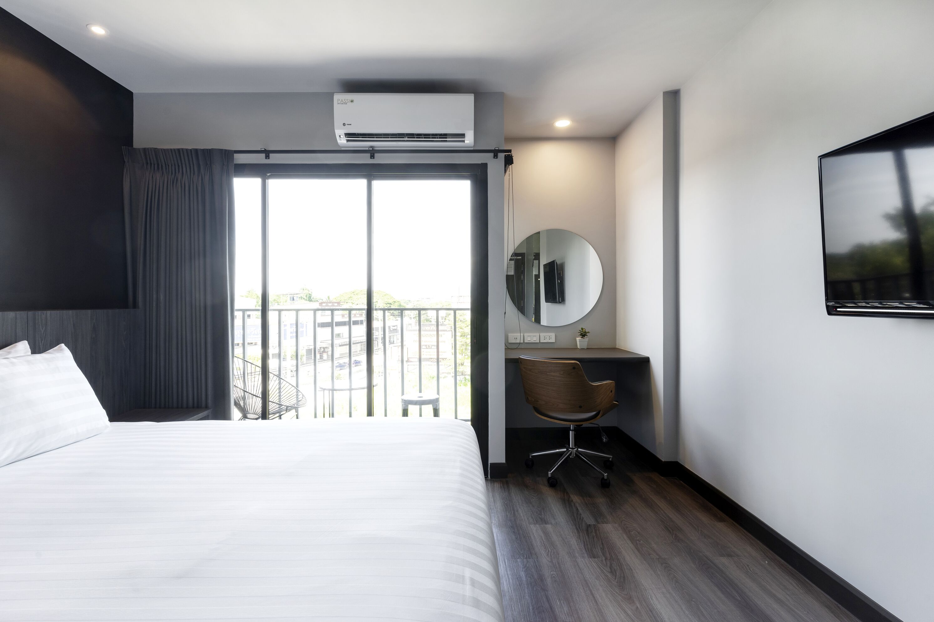 Ein modernes Hotelzimmer mit einem großen Bett, einem Schreibtisch mit Stuhl, einem runden Spiegel und Blick ins Freie durch ein großes Fenster.