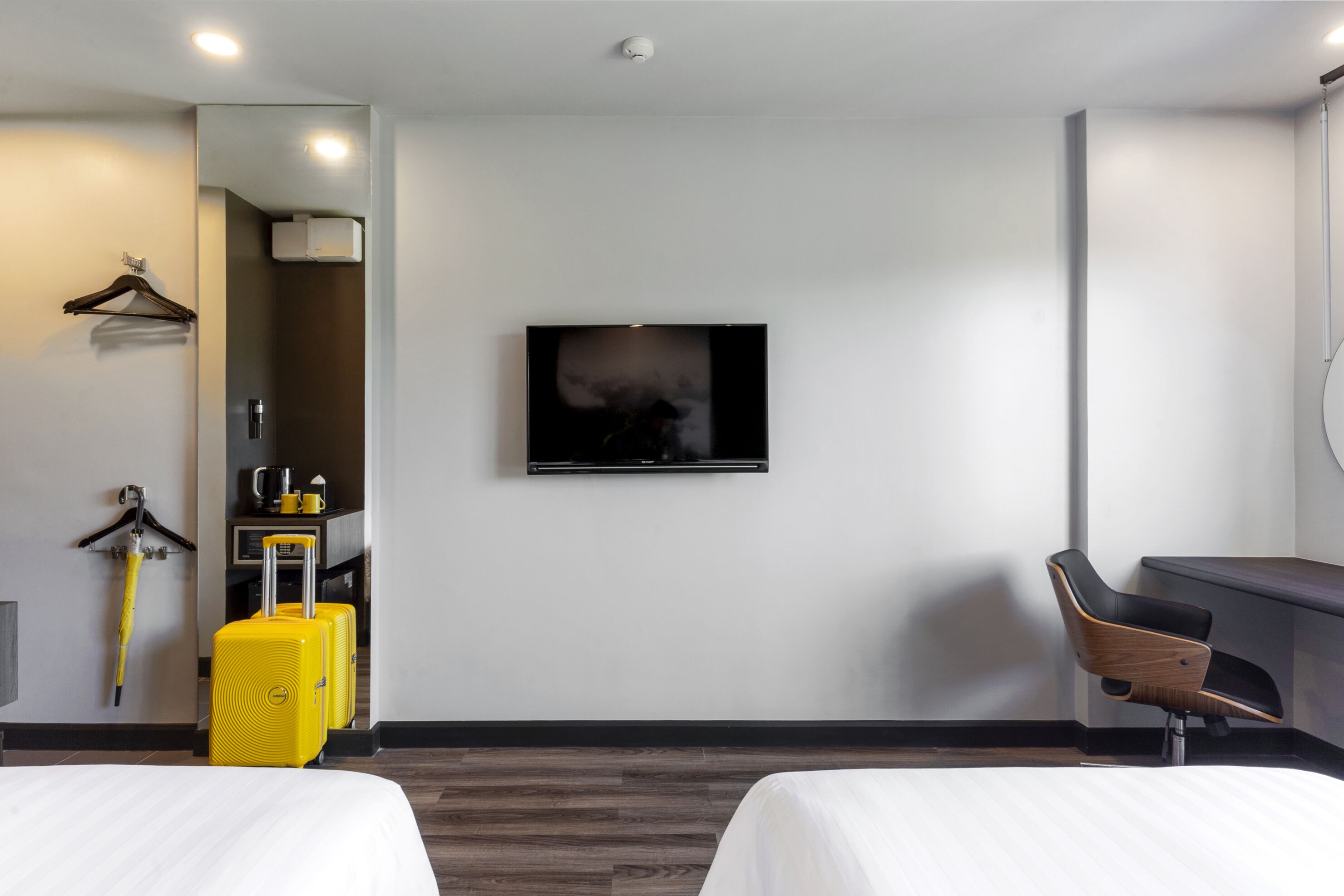 Une chambre d’hôtel avec deux lits, un téléviseur fixé au mur, un bureau, une chaise, un porte-manteau et une valise.