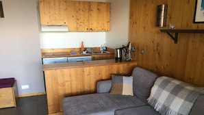 Ein Wohnzimmer mit grauem Sofa, einem Holztisch und einer Küchenzeile mit Holzausbau und Spüle.