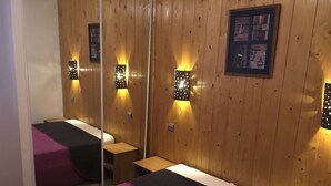 Ein Zimmer mit Holzvertäfelung, einem Bett mit violett-schwarzer Bettdecke, einem kleinen Holztisch und Wandleuchten.
