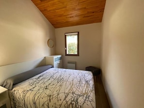 Une chambre avec un plafond en bois, un lit recouvert d’une couvre-lit à motifs, une table de chevet, une commode, un radiateur et une fenêtre donnant sur un paysage verdoyant.