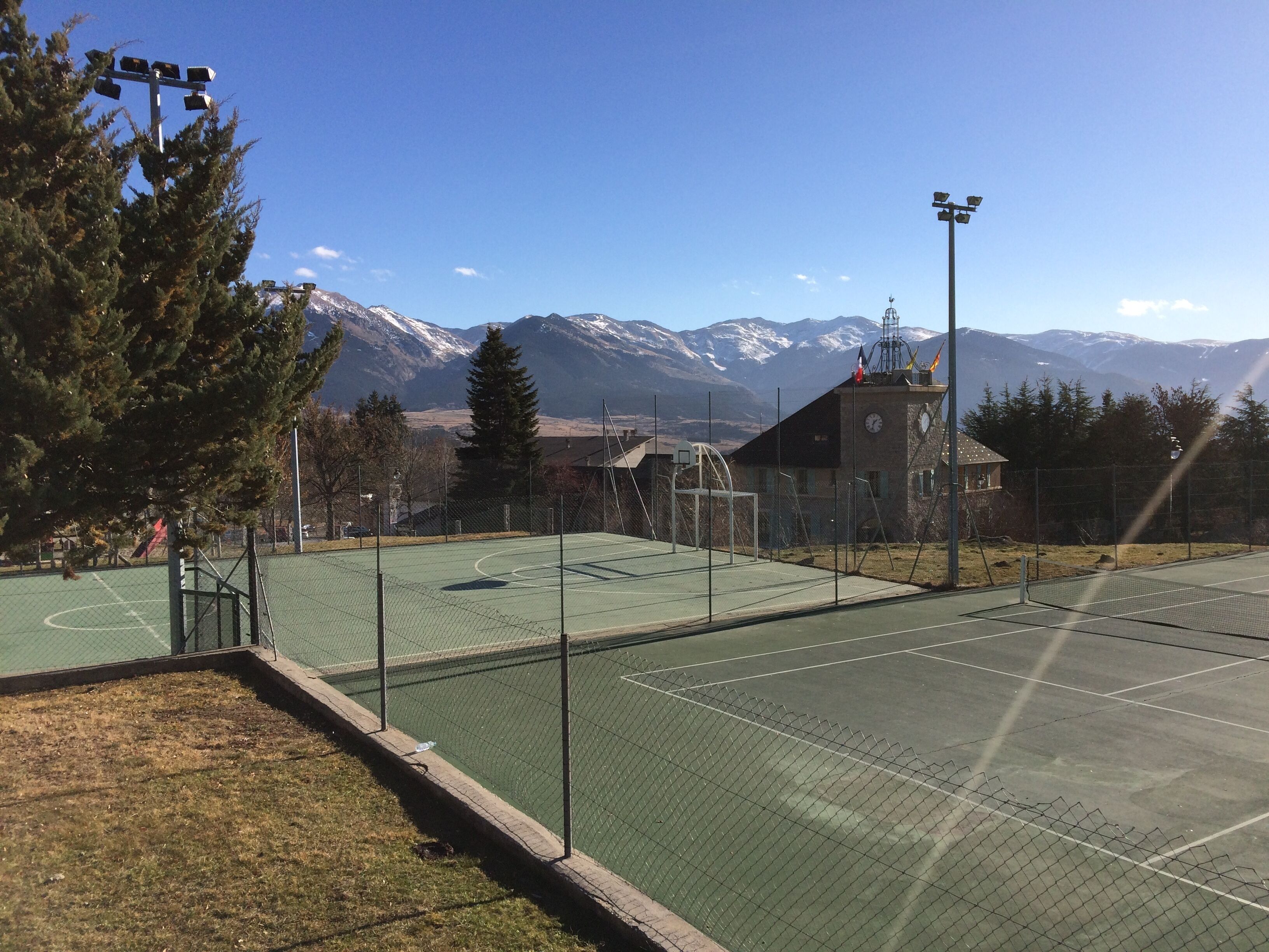 An outdoor tennis court with a net, surrounded by trees and a building with a clock tower.
