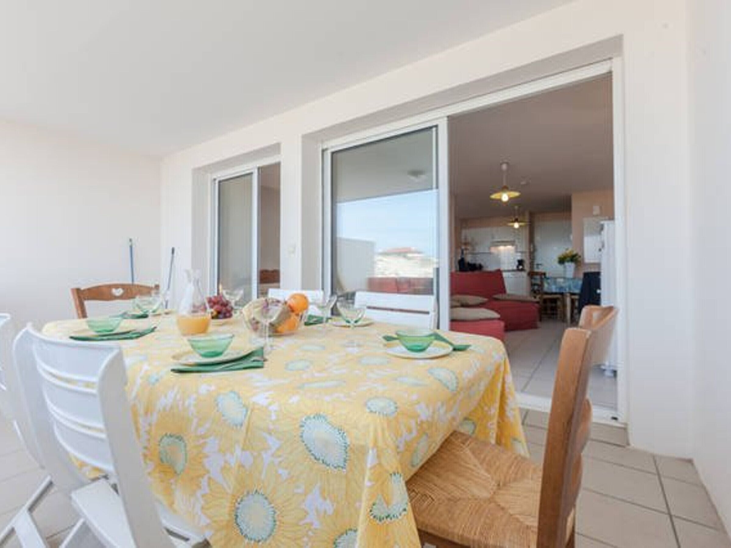 A dining area with a yellow tablecloth, white chairs, and a view of the kitchen and living area through glass doors.