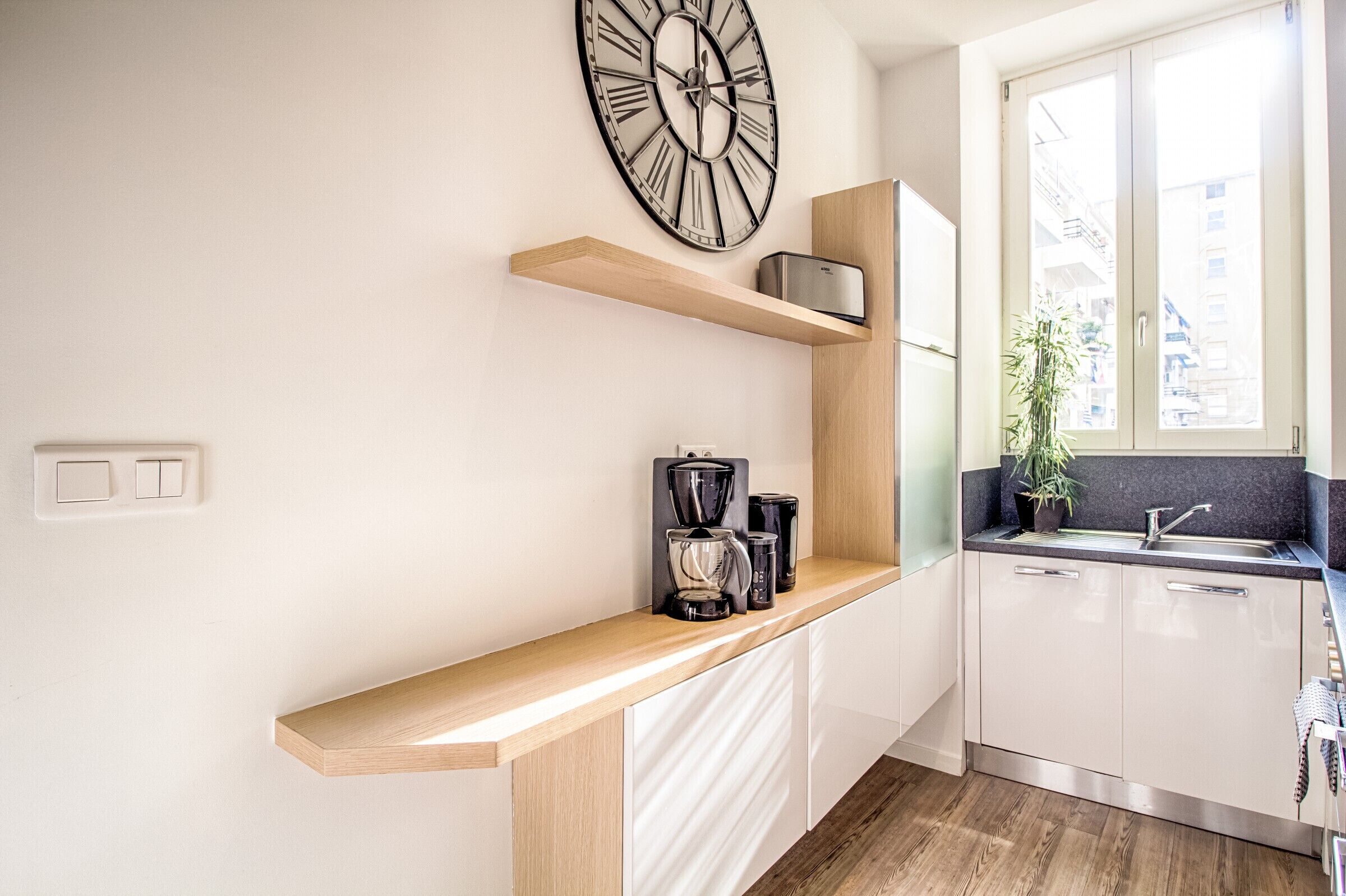 A modern kitchen with a wooden countertop, a coffee maker, a clock, and a window with a view of buildings.