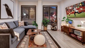 A modern living room with a grey sofa, a coffee table, and a large TV on the wall. There are two windows with blinds, a bookshelf, and a decorative globe. - The Sanctuary on Main Modern 1BR Walk to Town Lift (Park City)