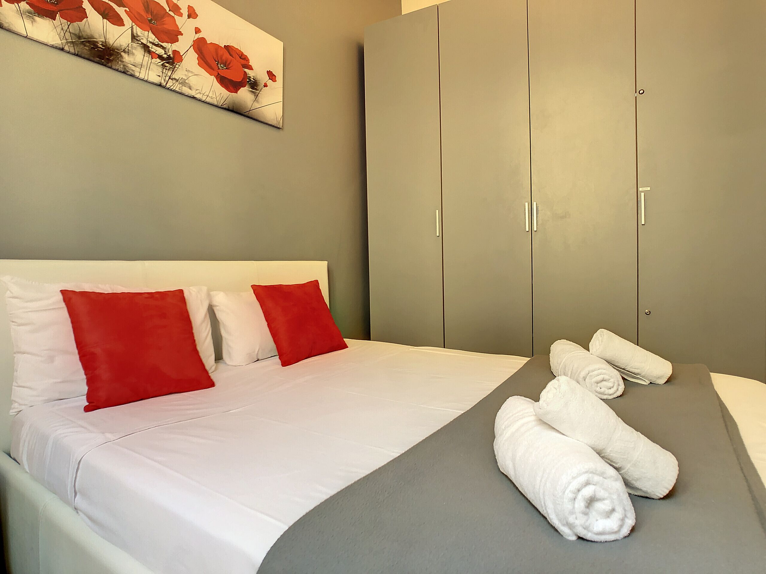 A neatly made bed with white and red pillows, a grey headboard, and a grey bed skirt. There are rolled white towels on the bed.