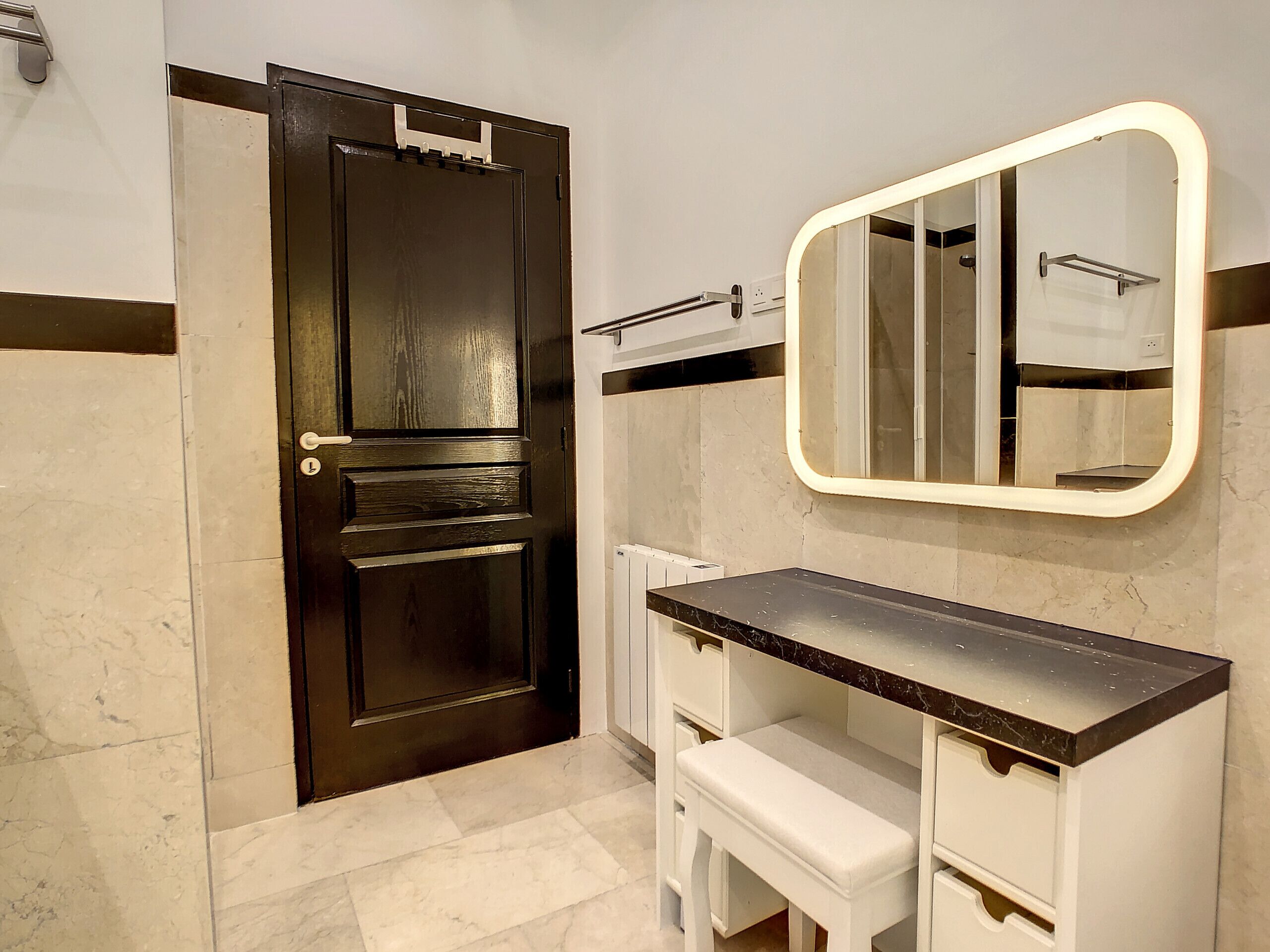 Baño con puerta de madera oscura, tocador blanco con encimera negra, un banco y espejo con marco iluminado.