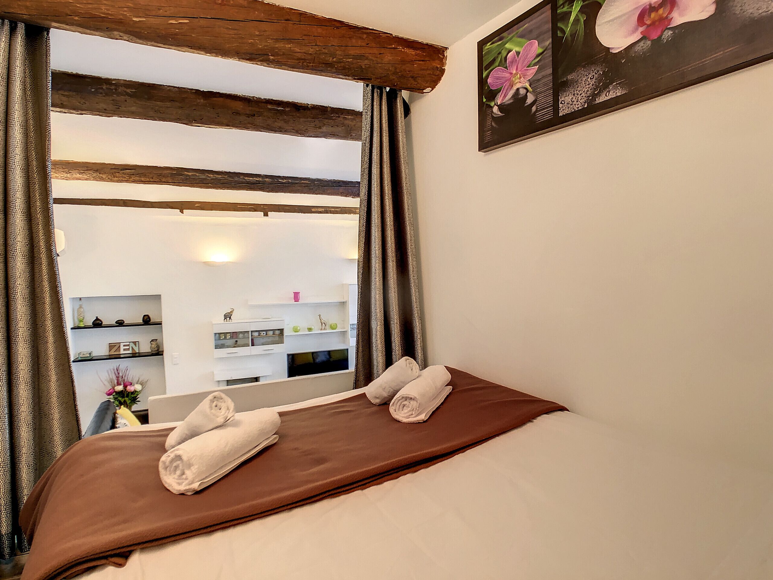 A room with a bed, towels, a wooden ceiling, a wall-mounted picture, and a view of a bathroom with shelves and a mirror.