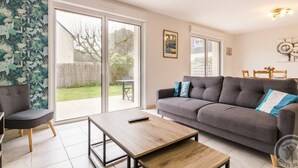 A living room with a grey sofa, a wooden coffee table, and a chair. There is a sliding glass door leading to a patio with a table and chairs.