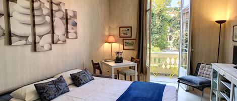 A hotel room with a bed, a desk, a chair, and a balcony with a view of trees.
