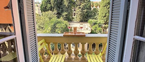 En balkon med gule stole og et bord, der ud til en have med træer og en bygning i baggrunden.