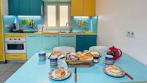 Une cuisine compacte aux armoires turquoise et jaunes, avec un petit four et une table à manger dressée avec des assiettes, des tasses et un panier de pain.