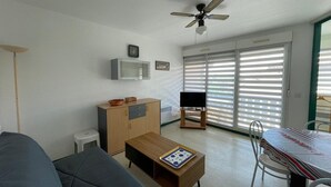 A compact living space with a sofa, a wooden coffee table, a television, a ceiling fan, a microwave, and a dining table with chairs.