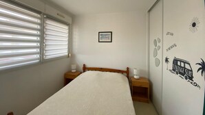 A bedroom with a bed, a window with blinds, a bedside table, a nightstand, a wall with a sun and palm tree decal, and a framed picture on the wall.