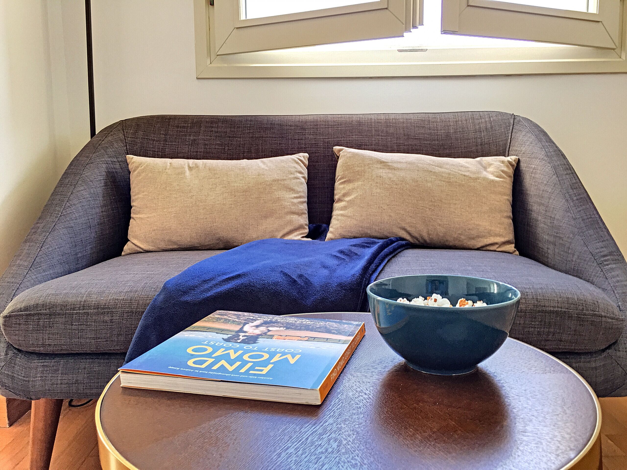A grey sofa with two beige cushions, a blue throw blanket, a book titled 'Find Mom and Dad', and a blue bowl with popcorn on a round wooden table.