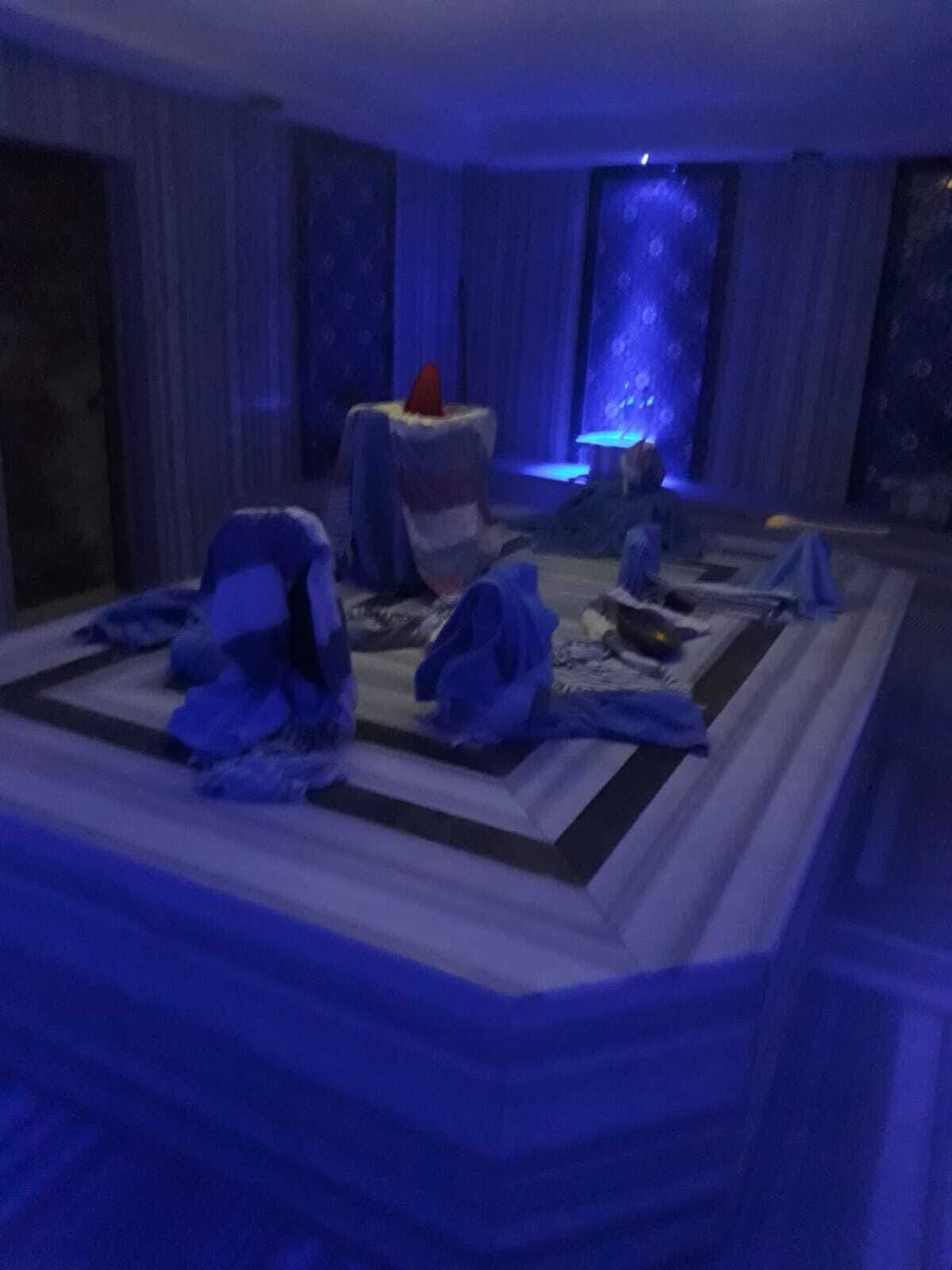 Ein Zimmer mit einem großen, gemusterten Teppich, einem kleinen Tisch mit einem roten Gegenstand und einem blau beleuchteten Hintergrund.