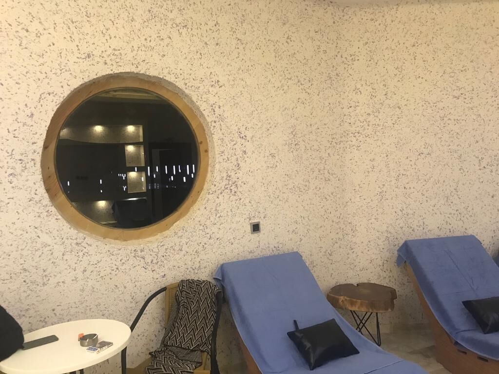 Uma sala de spa com um espelho redondo, duas macas de massagem, uma mesinha com uma cadeira e uma poltrona estampada.