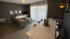 A modern living room with a sofa, a dining table, and a TV. - Garden-Level Apartment 100m from Beach with Parking & Terrace (Capbreton)