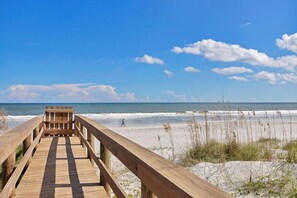 Ein hölzerner Steg führt zu einem Strand mit blauem Himmel und vereinzelten Wolken.