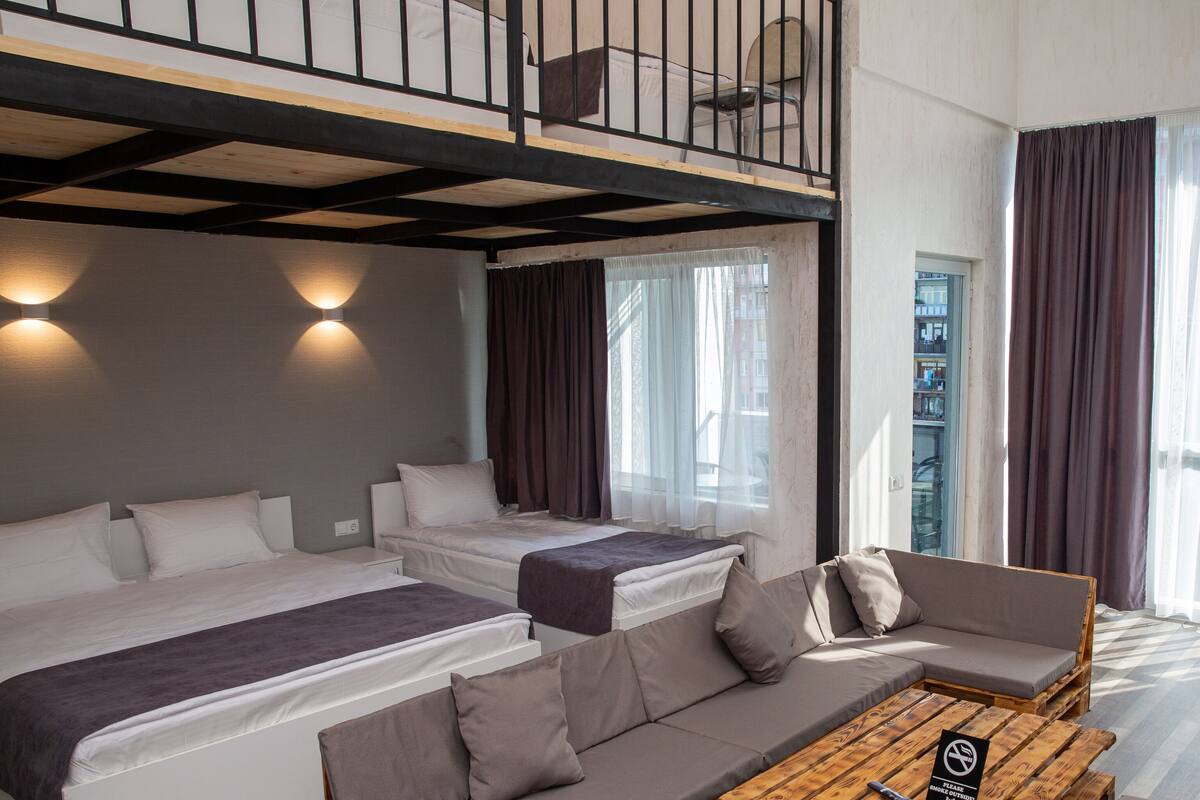 A modern hotel room with a loft bed, a sofa, a coffee table, and large windows with curtains.