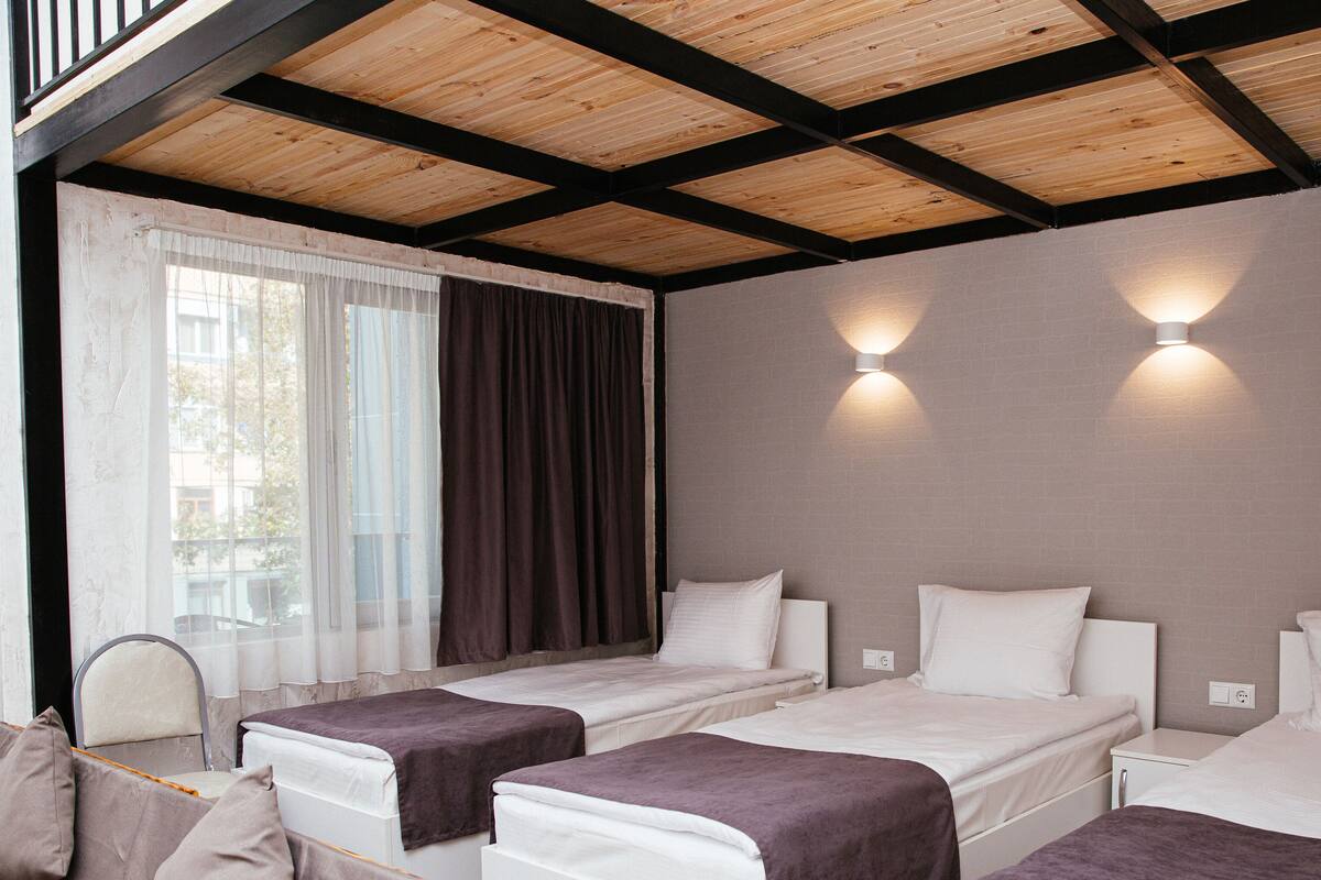 A hotel room with two single beds, a window with curtains, and a wooden ceiling.