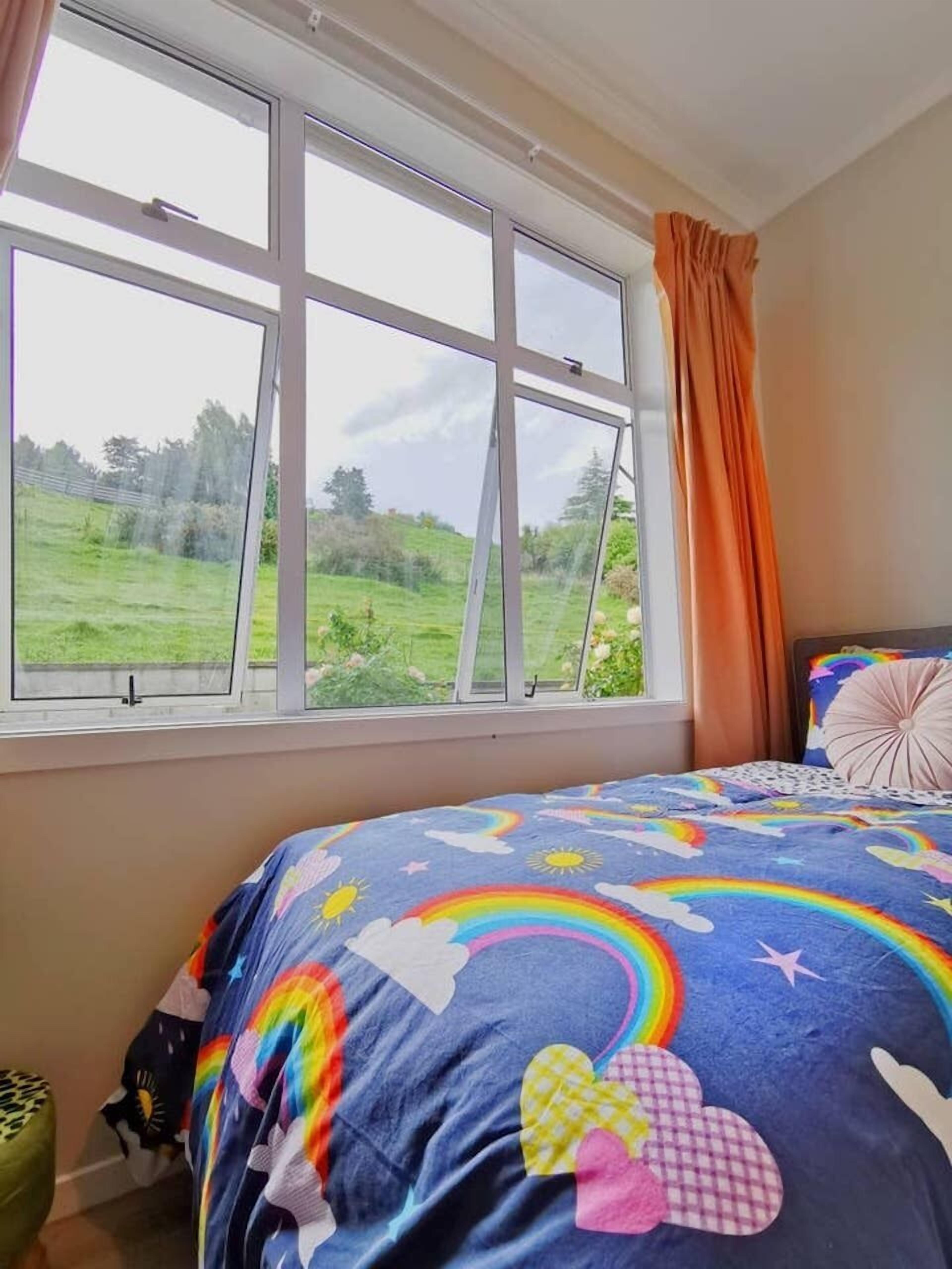 Een slaapkamer met een groot raam, een bed met een kleurrijk regenboog- en wolkbed, en uitzicht op een groen veld.