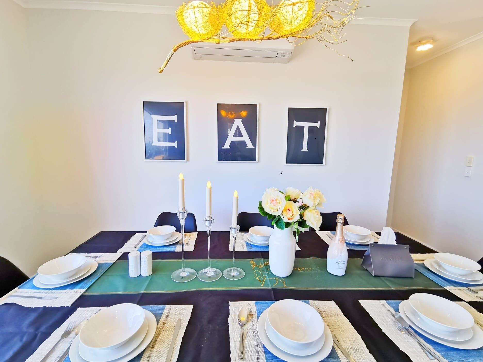 A dining table set with plates, candles, and a vase of flowers, with "EAT" letters on the wall above.