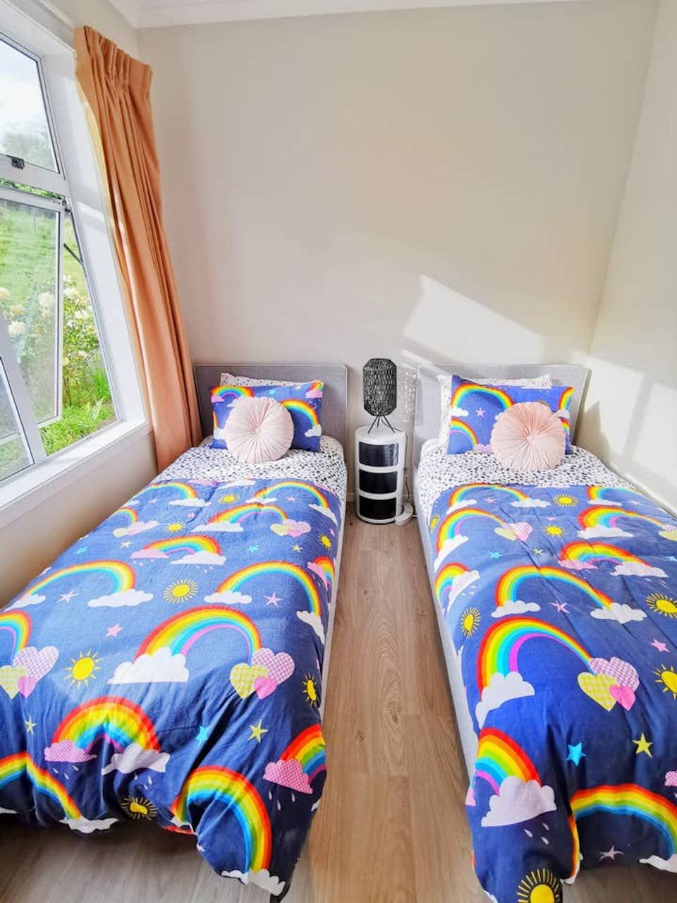 Quarto com duas camas, cada uma com colcha de retalho colorida e estampa de nuvens. Um criado-mudo com luminária fica entre as camas.