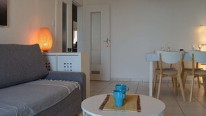En moderne stue med en grå sofa, et rundt sofabord med en blå kop, et hvidt spisebord med stole, og en tændt lampe.