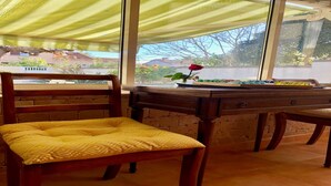 Ein Zimmer mit einem Holzsessel, einem Tisch mit Blumen in einer Vase und Blick durch große Fenster.