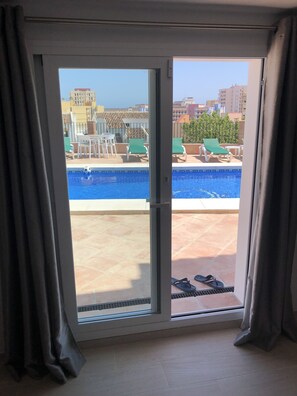 A view through a sliding glass door to a rooftop pool area with lounge chairs and a swimming pool. - Villa Chispita (Fuengirola)