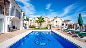 A swimming pool with a blue mosaic design, surrounded by lounge chairs and umbrellas, with buildings in the background. - Villa Chispita (Fuengirola)